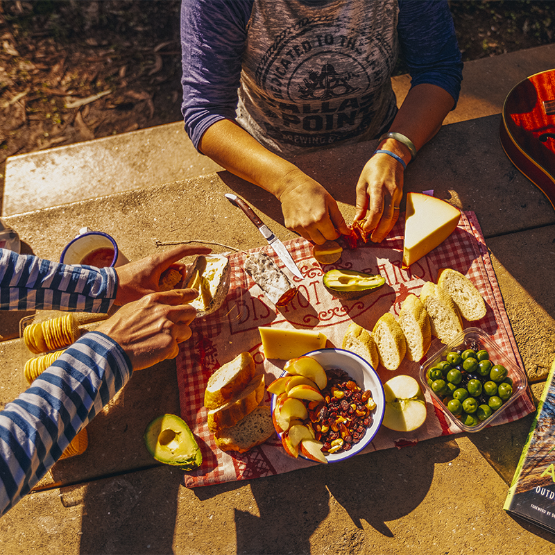 A couple sitting at a picnic table at a campsite having some snacks and coffee in the sun.