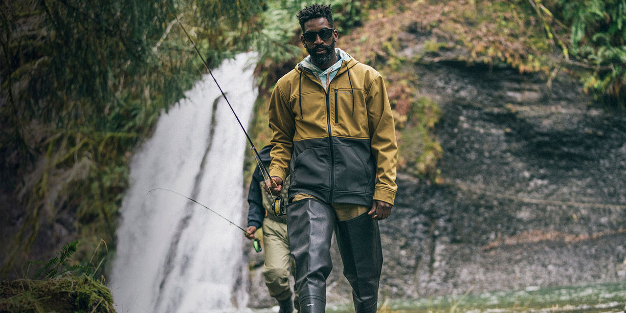 Fishermen walk with fishing poles along a river with a waterfall behind him