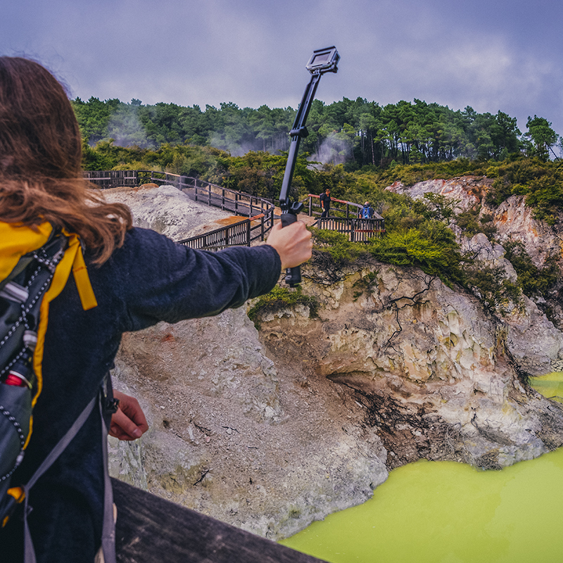 A woman holds a POV camera (GoPro) on a stick over the Devil's Bath at Waiotapu Geothermal Wonderland, a colorful volcanic area in New Zealand.