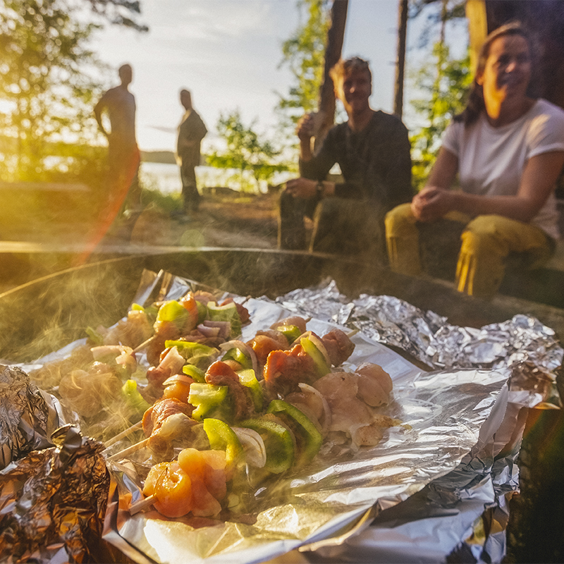 A group of people gather around food on skewers being grilled In a firepit 