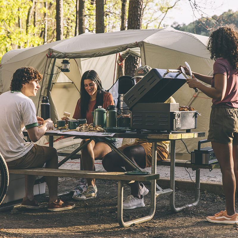 Friends sit at a campsite bench while they cook