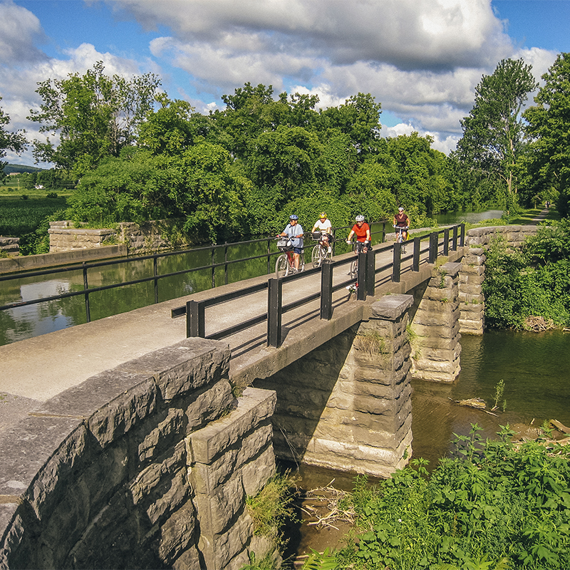 A paved, stone overpass over the canal with several bicyclists on the path of  the Erie Canalway Trail.