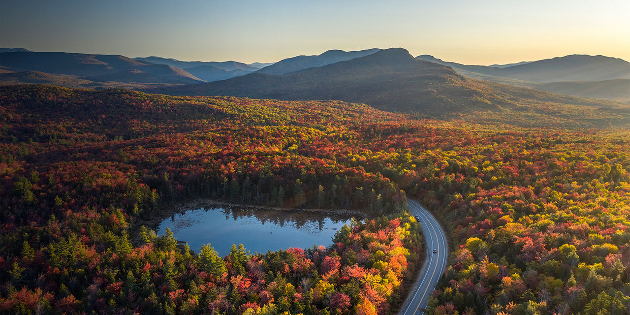 A road, highway through a forest with trees in fall colors of red, orange and yellow, lake, mountains, Kancamagus Highway, White Mountains, New Hampshier