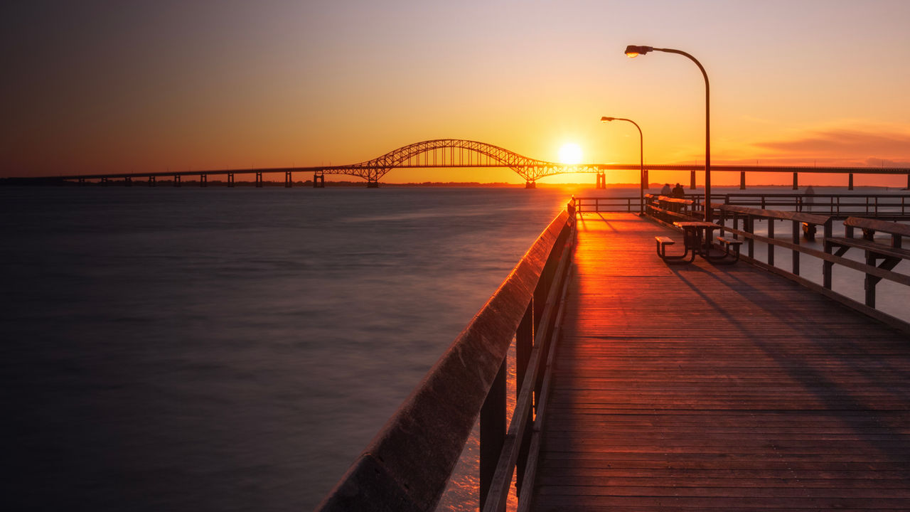 Wooden fishing pier leading towards an arched bridge, as the sunset dips below the horizon. Captree State Park - Long Island New York