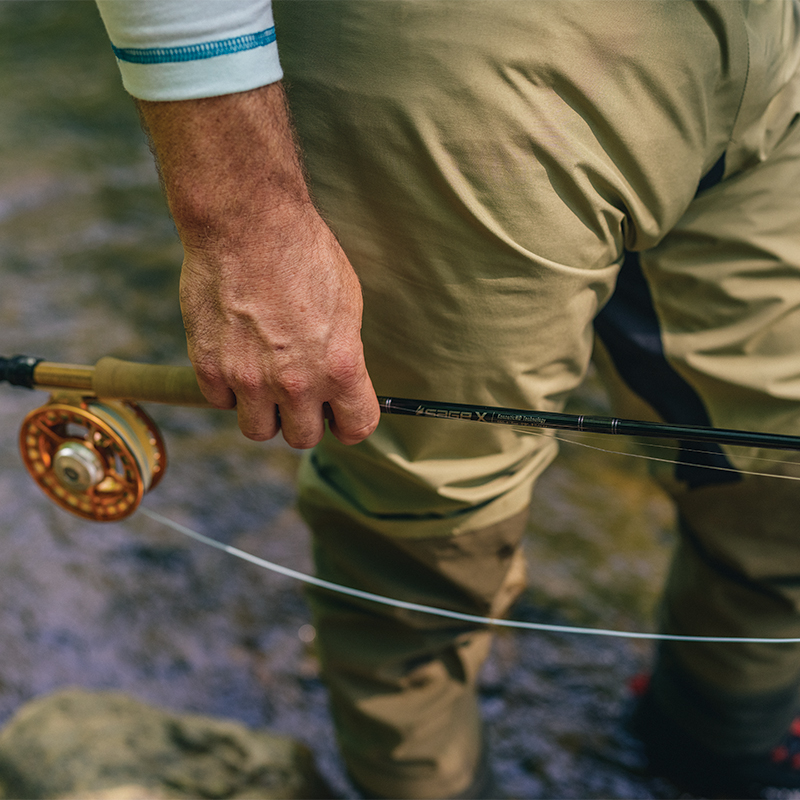A man standing in a creek, wearing waders and holding a fishing rod.