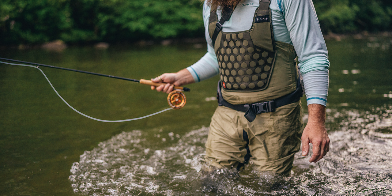 A man standing, wearing waders, while fishing in a creek.