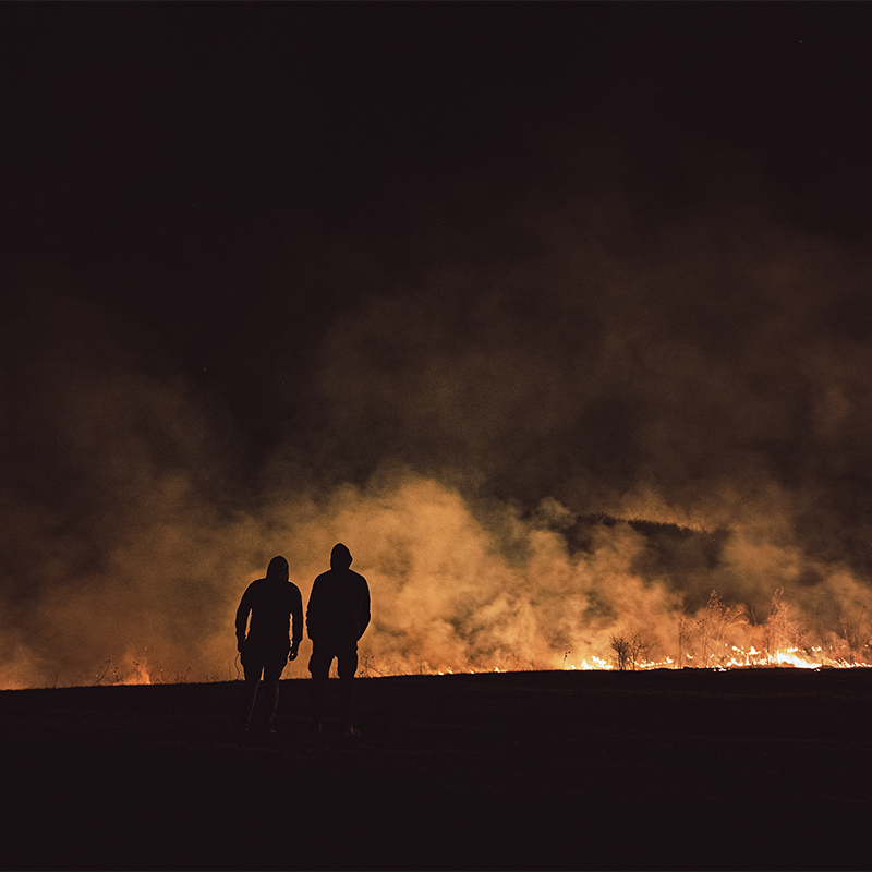 Two people walking with a view of wildfire in a field in the evening