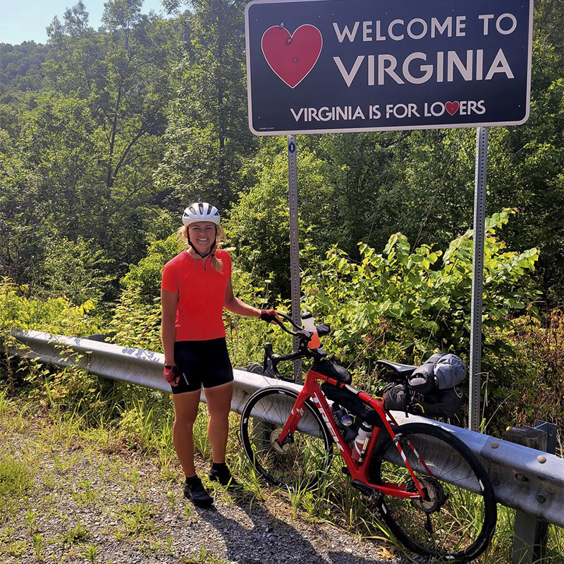 Brooke Barney poses with her bike in front of a Welcome to Virgina sign.