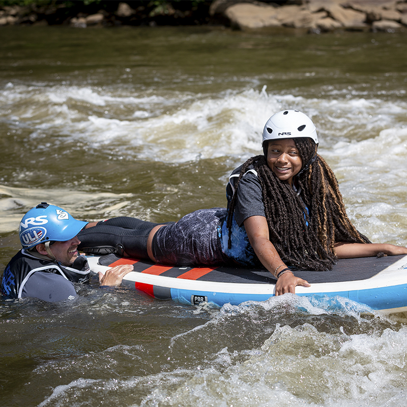 A girl hangs on to a paddle board with a guide as part of First Waves