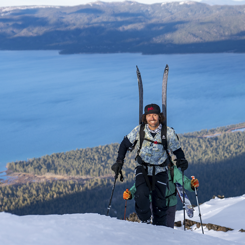 Jeremy Jones smiles for the camera hiking up a mountain with skis