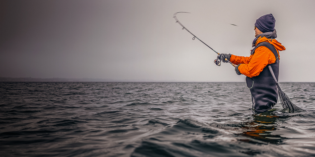 Angler in Wathose mit Spinnrute und Kescher beim Wurf mit Köder im Winter im kalten Meer an der Küste bei Wolken, Angler in Wathose mit Spinnrute und Kescher beim Wurf mit Köder