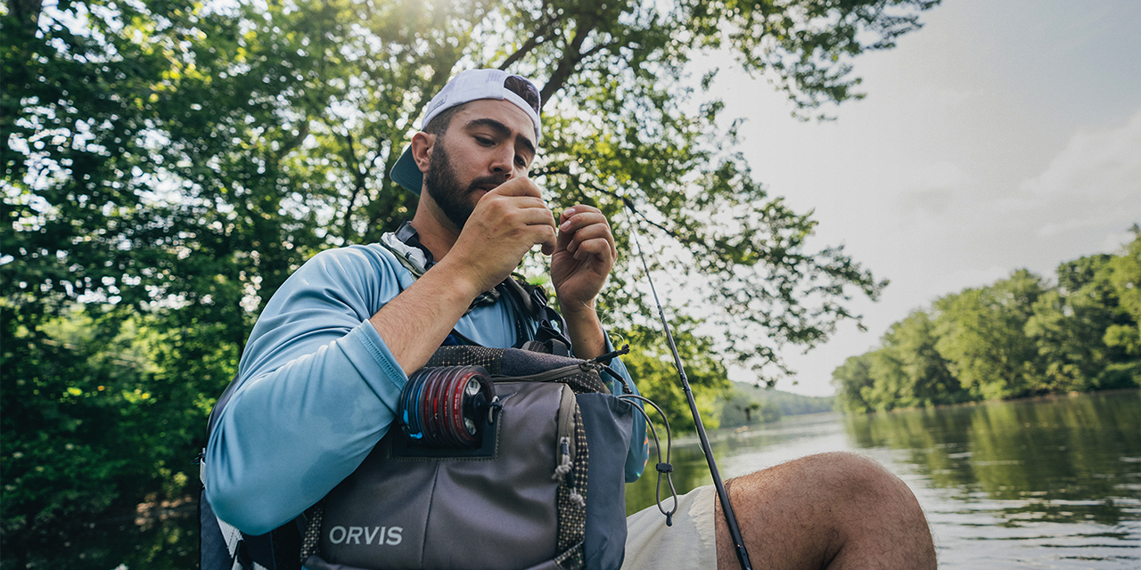 A man holding a lure, wearing a light blue long sleeve shirt, white hat with a fishing bag in his lap.