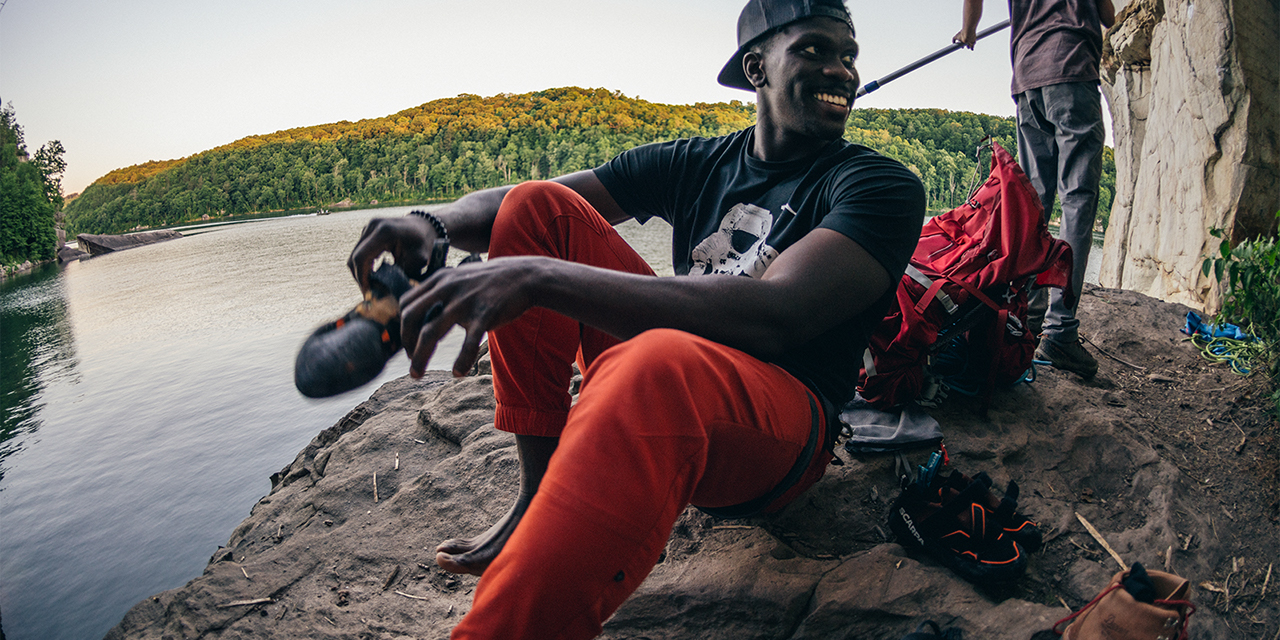 A man sitting and smiling on a rock, wearing a black shirt and orange pants with someone stanindg behind him with a river and forest behind them.