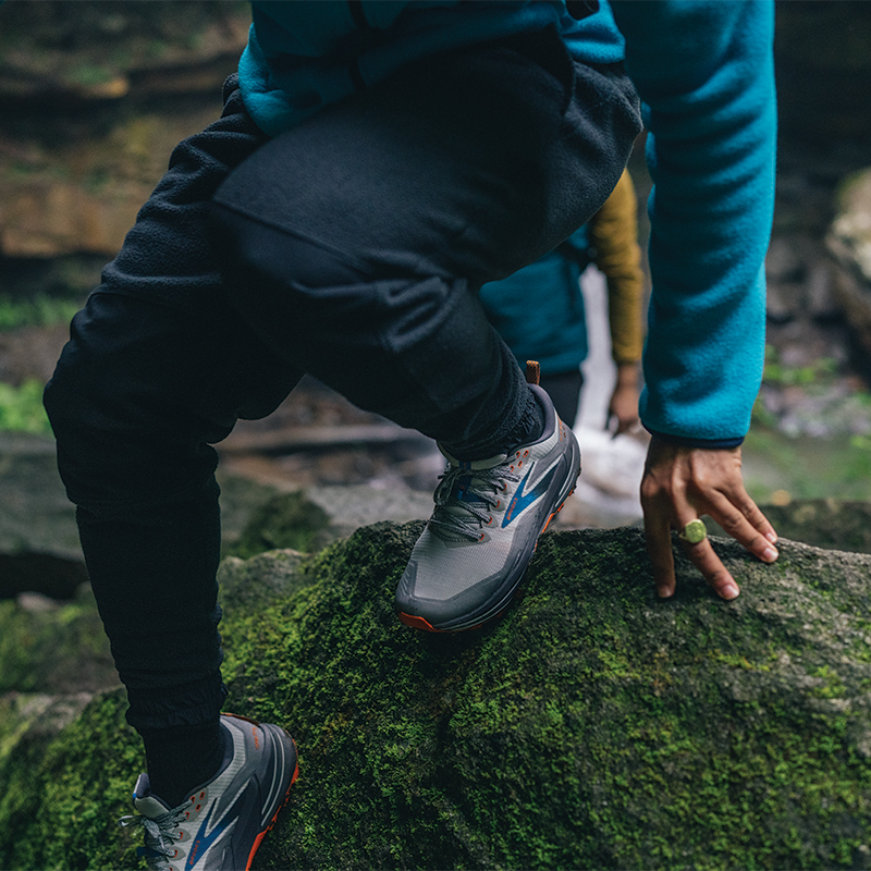 A man climbing over a rock , he is wearing a light blue full zip fleece, navy blue pants and grey shoes.