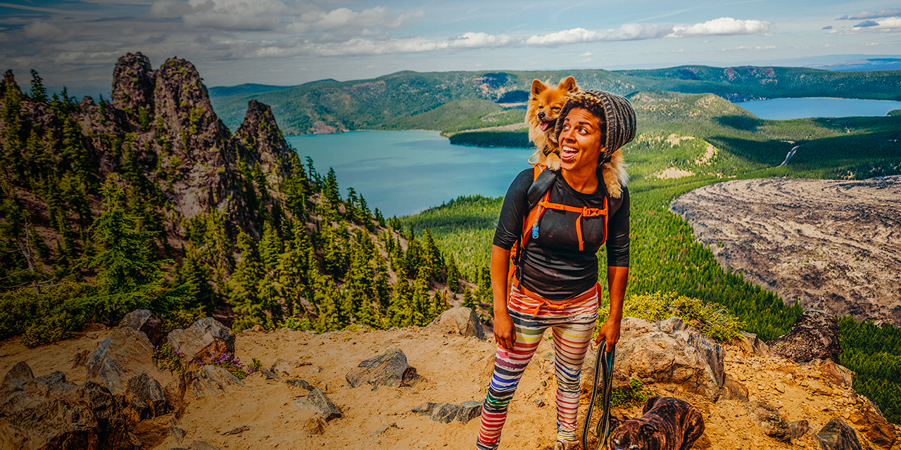A woman hikes with her dog in a backpack and on a leash