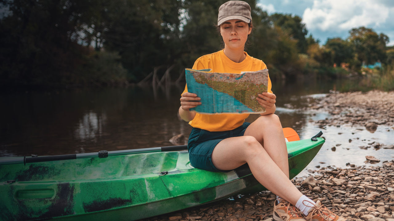 A young woman is sitting on a kayak, holding a paper map to check the route. 