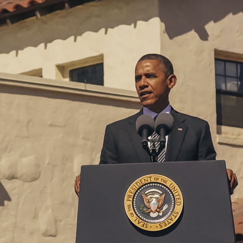 President Barack Obama speaks at a podium in front of a white stucco building