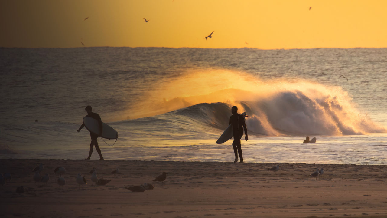 Silhouette of a surfers walking on the beach at sunset, with waves crashing in the background. Long Beach New York