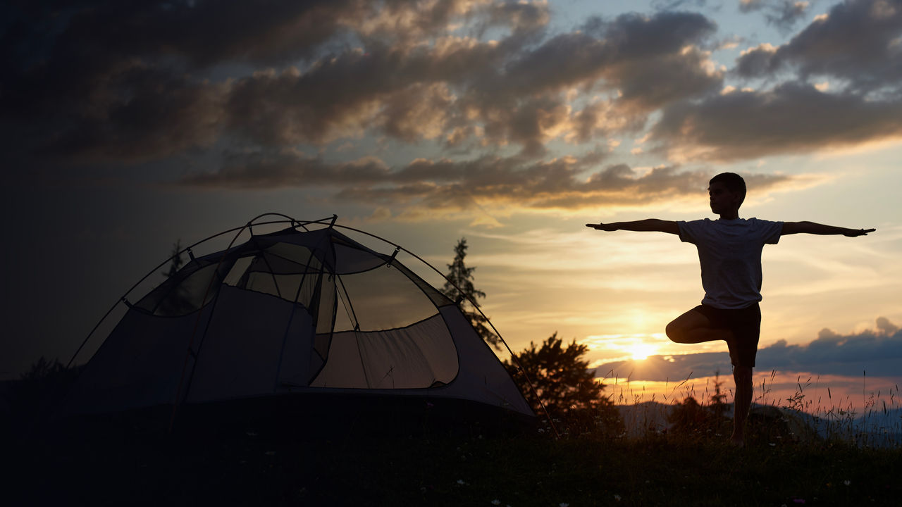 Silhouette of boy at practice yoga on the top of mountain at the dawn near campsite under sky with clouds and bright morning sun.