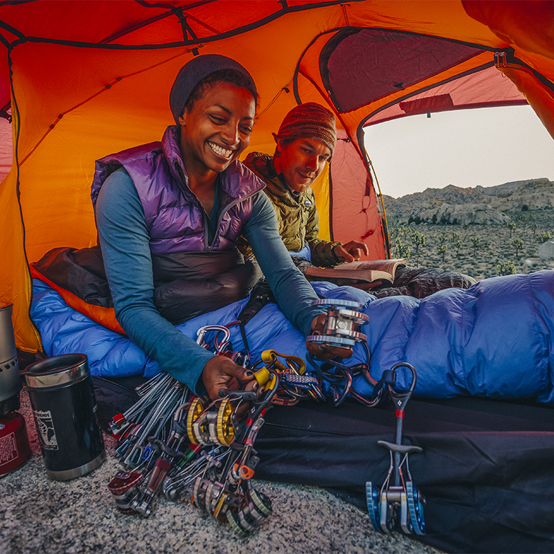 A woman and a man prepare coffee, sort gear and look through the local climbing guide book in their tent as morning breaks in Joshua Tree National Park - California