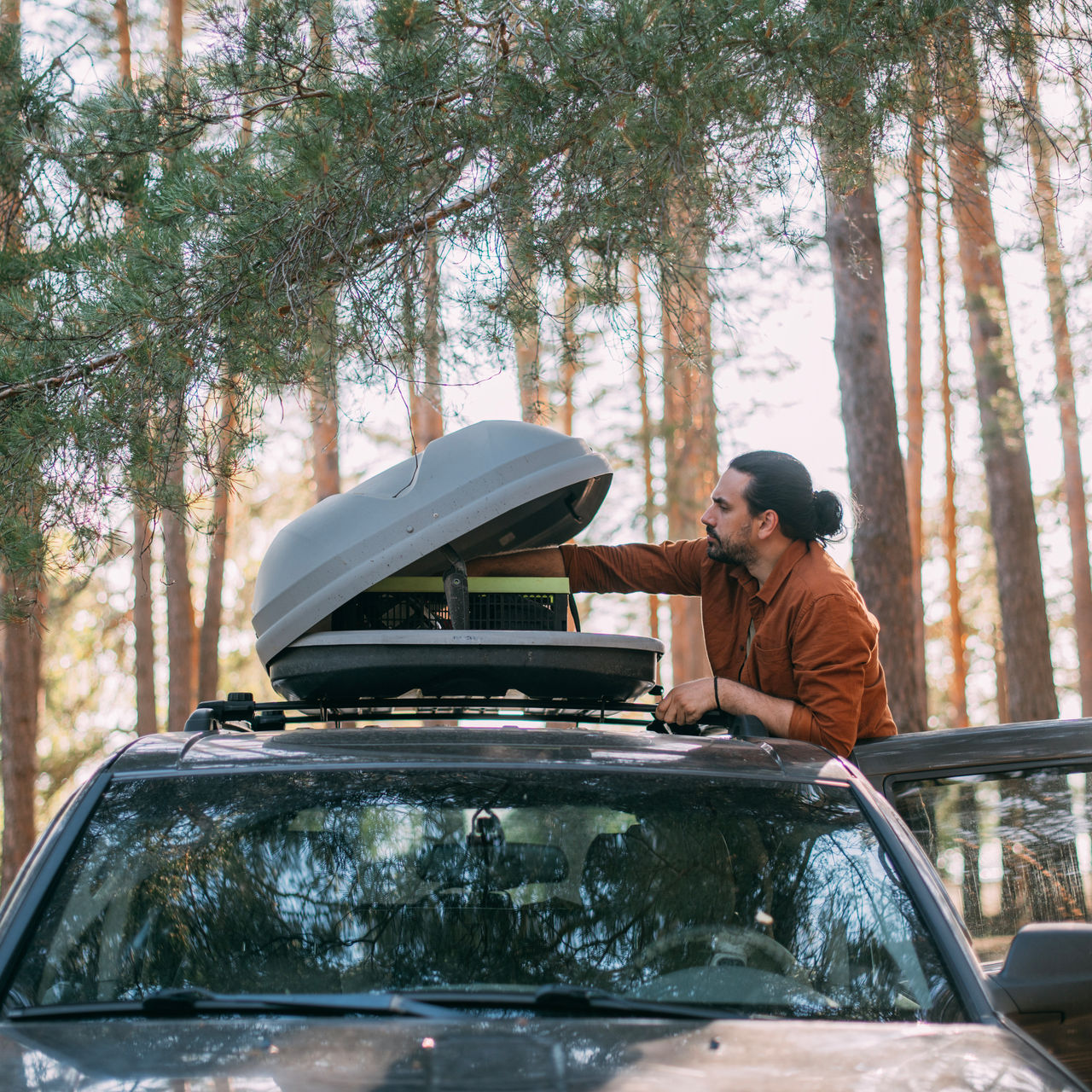 A young man opens the roof rack of a car in a pine forest on the shore of a lake at sunset.